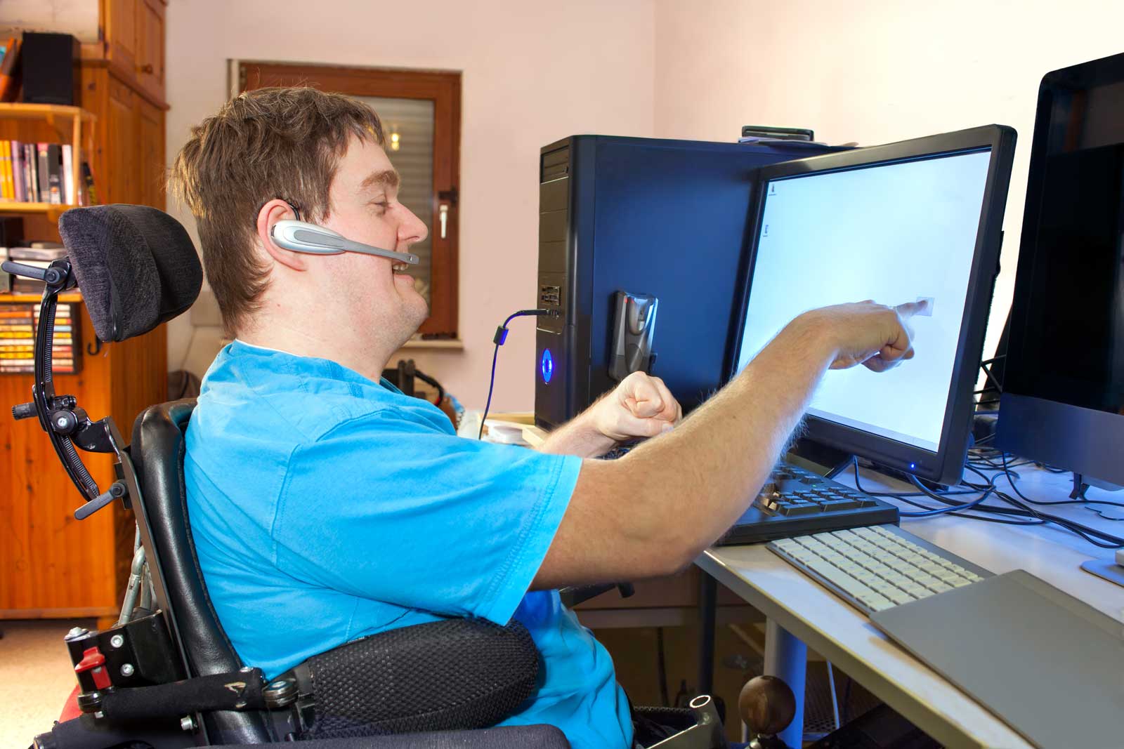 A young man with infantile cerebral palsy caused by a complicated birth sitting in a multifunctional wheelchair using a computer with a wireless headset reaching out to touch the touch screen
