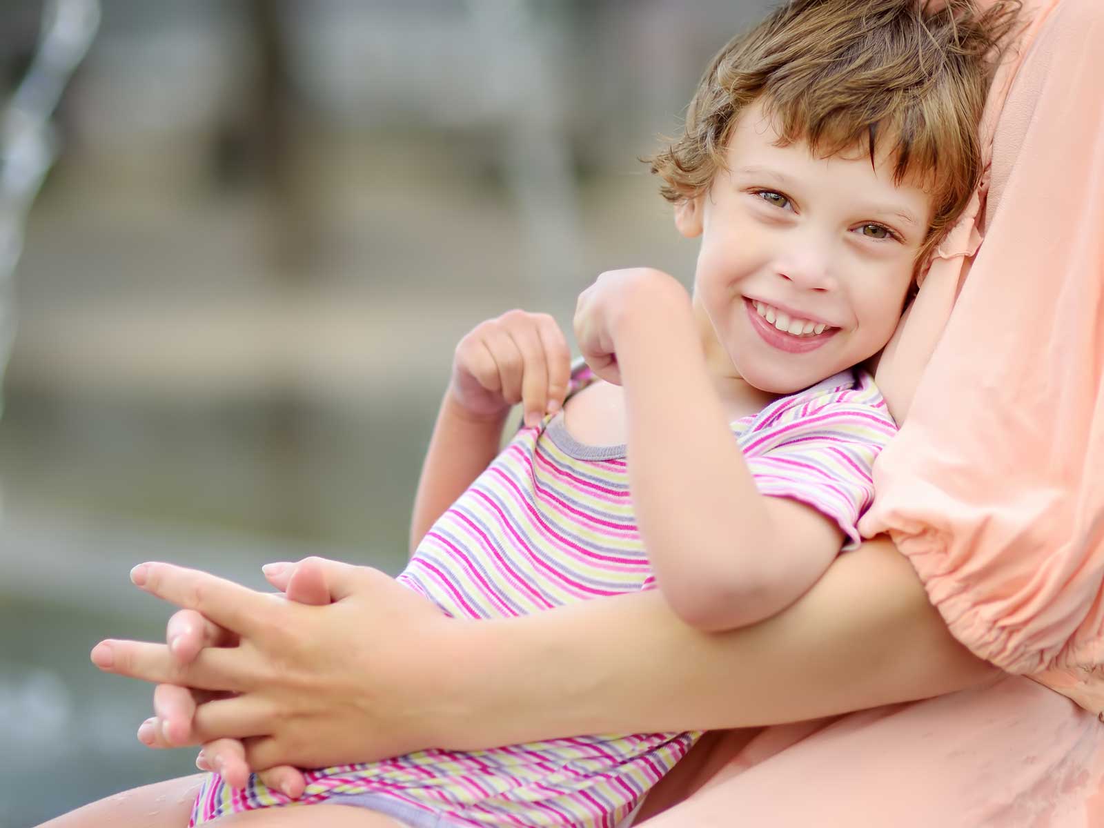 lose-up portrait of beautiful disabled girl in the arms of his mother having fun in fountain of public park at sunny summer day.