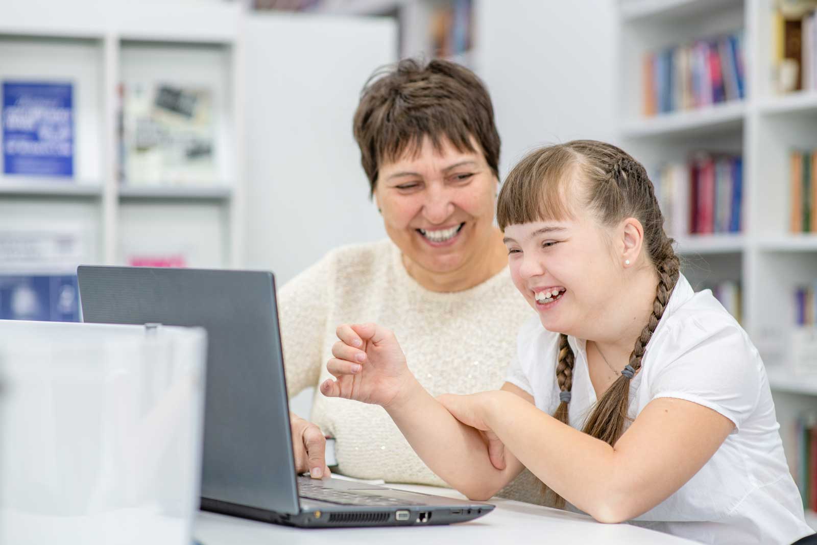 Smiling girl with down syndrome is uses a laptop with her teacher at library. Education for disabled children concept