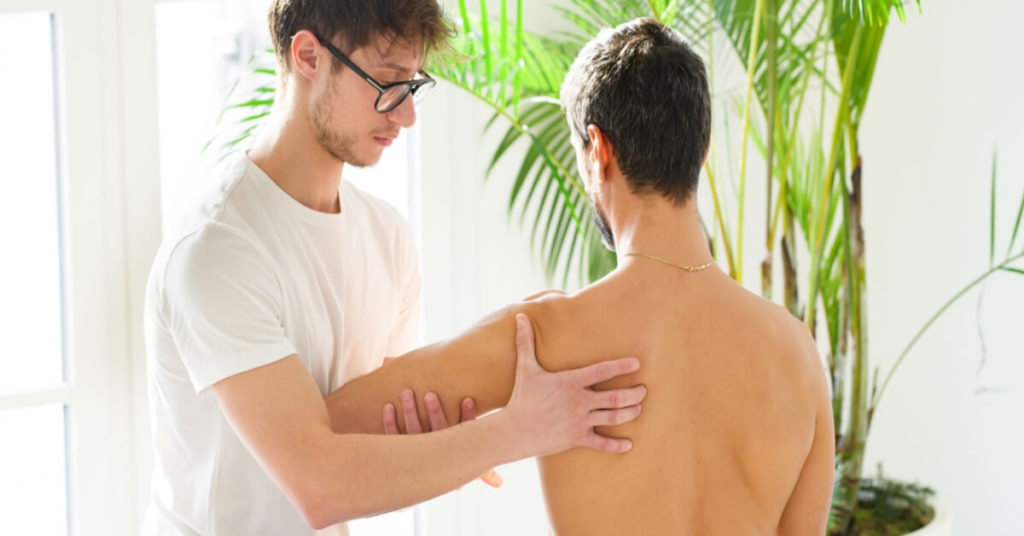 Osteopath assessing the scapula, shoulder blade, wing bone or blade bone of a young man in his clinic in a close up high key view