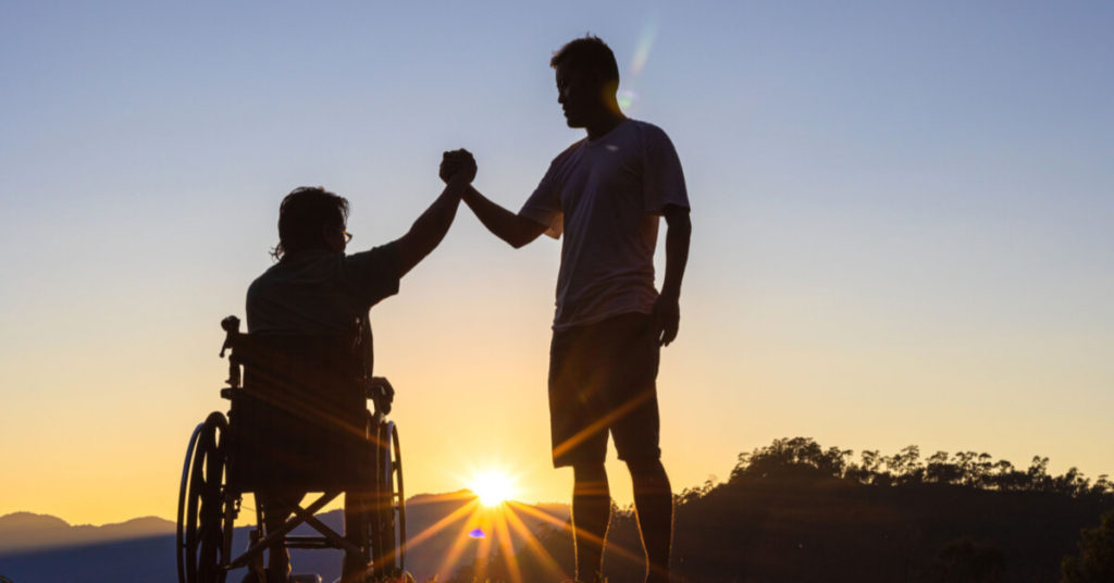 Silhouette of joyful disabled man in wheelchair raised hands with friend at sunset