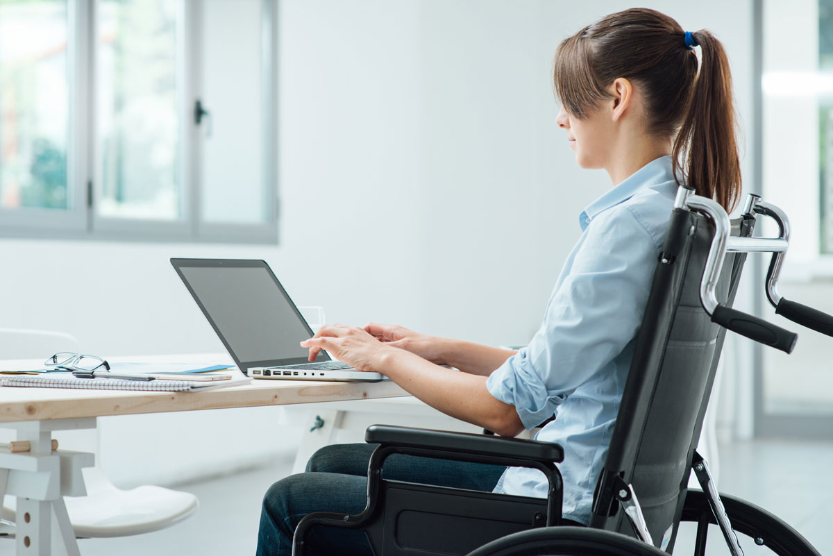 Young disabled business woman in wheelchair working at office desk and typing on a laptop